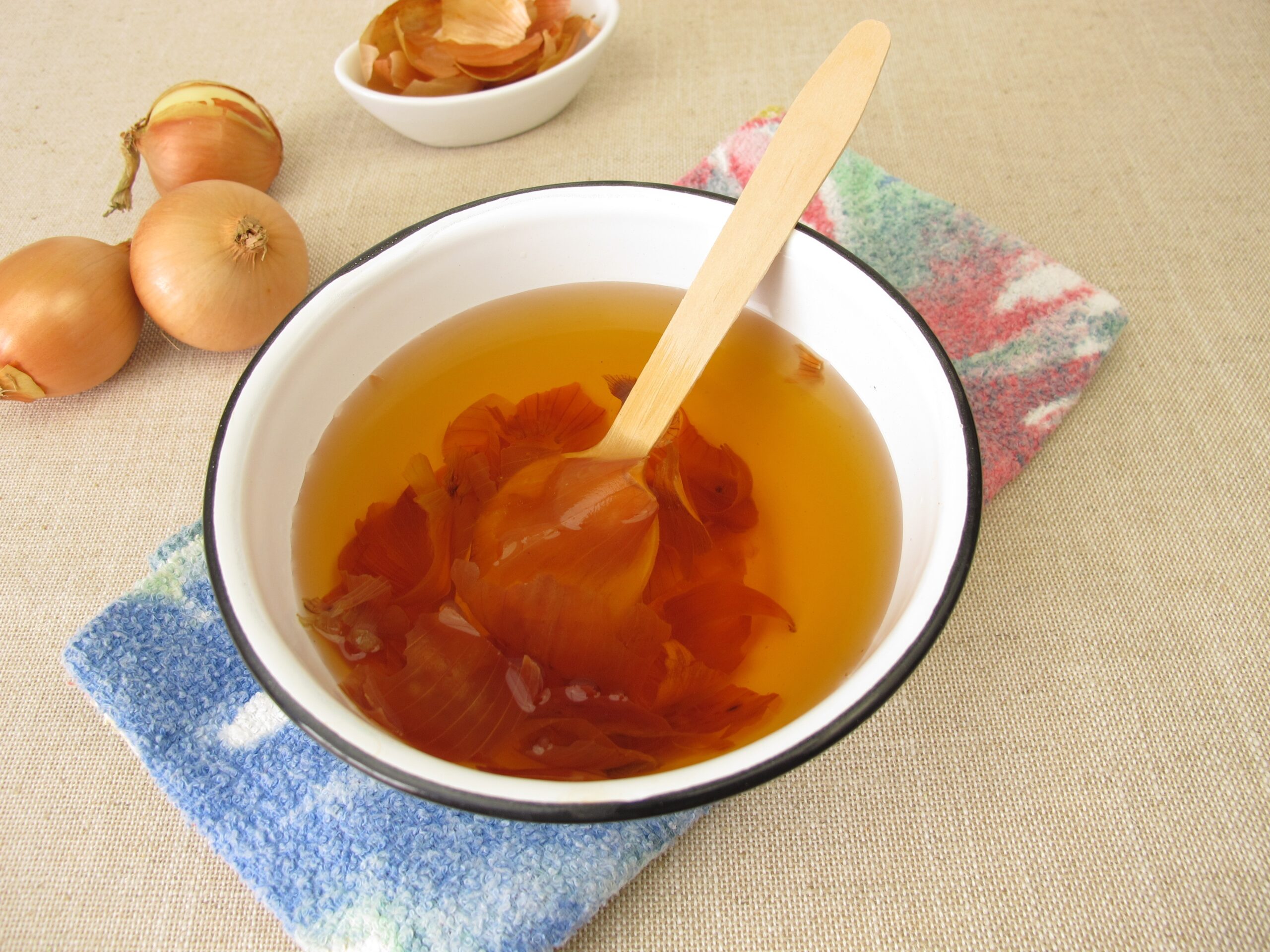 Un castron cu apa, frunze de ceapa, o lingura de lemn, pe servetel, cu ceapă si castron cu frunze de ceapa in spate